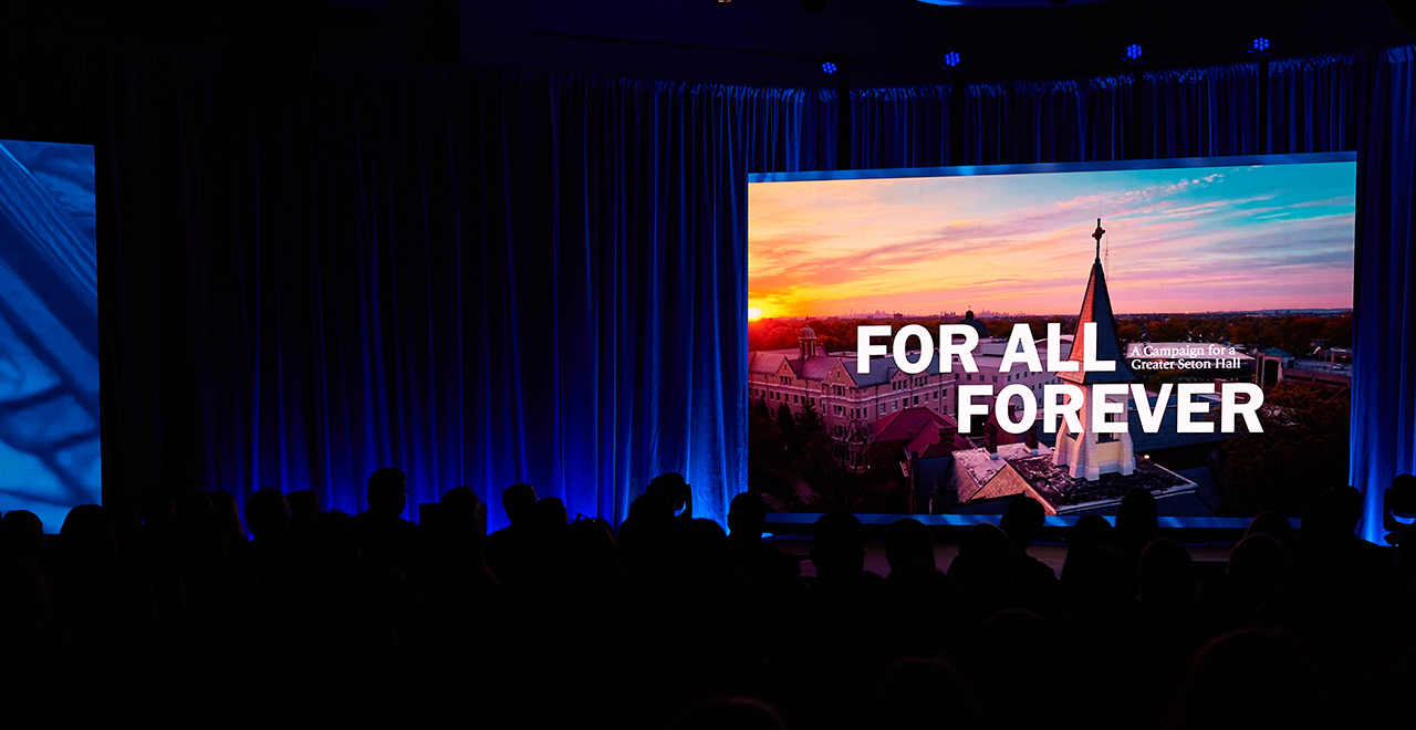 A photo of a screen that reads For All, Forever.